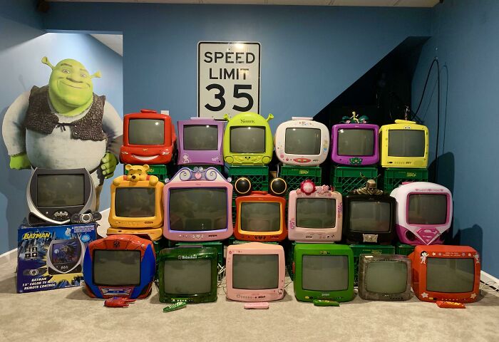 Collection of colorful vintage TVs stacked, with a Shrek cutout and a speed limit sign in the background.