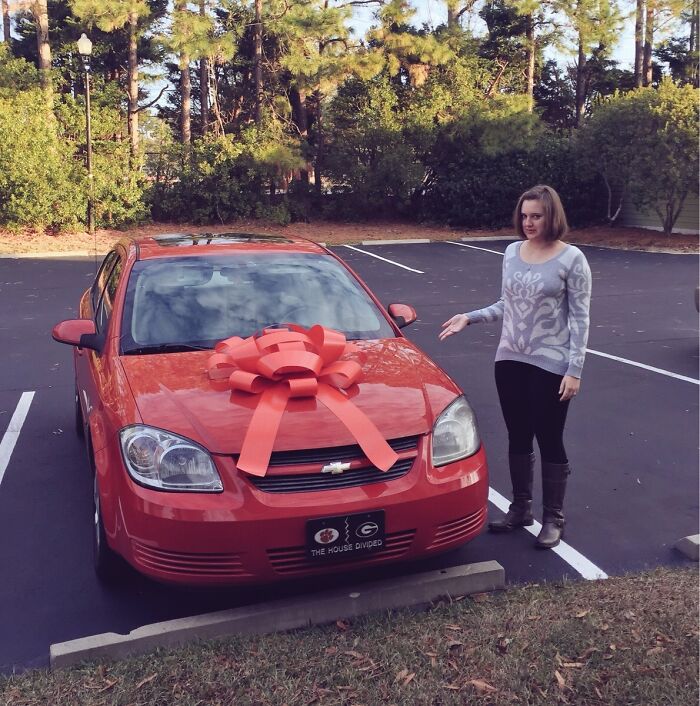 A woman stands beside a red car with a large bow, showcasing a hilarious trolling Christmas gift.