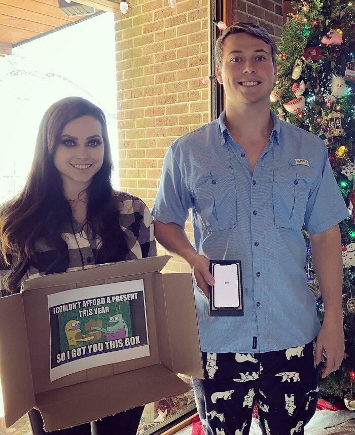 Two people exchanging hilarious trolling Christmas gifts, including a box and an empty phone case, by a decorated tree.