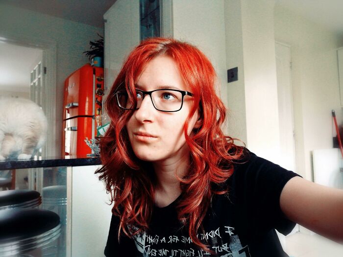 Person with red hair and glasses looking thoughtful, with a cozy kitchen in the background, representing wholesome vibes.