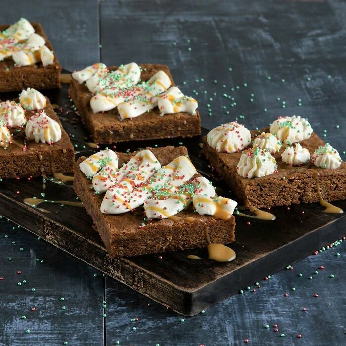 Festive brownies topped with cream and colorful sprinkles, perfect Christmas snack food idea on a rustic wooden board.