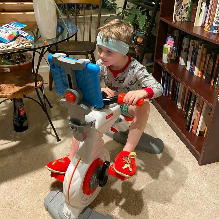 Child on a toy exercise bike with tablet, wearing Christmas pajamas, demonstrating hilarious trolling Christmas gifts.