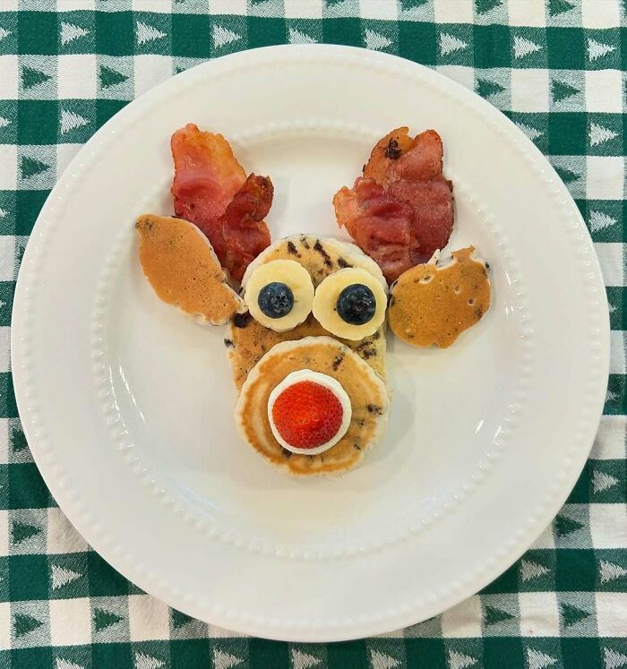 Reindeer-shaped Christmas snack made with pancakes, bacon, banana, blueberries, and a strawberry on a white plate.