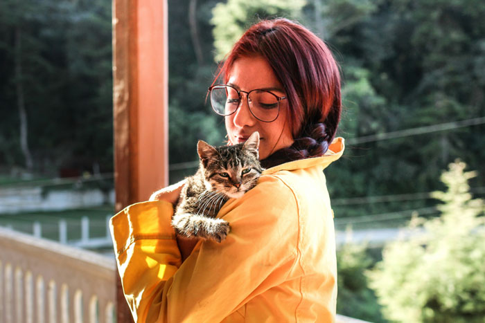 New mom holding a cat outdoors, wearing glasses and a yellow jacket, surrounded by greenery.