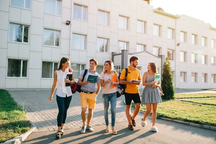 Group of young adults walking and talking in front of a building, carrying books and backpacks, romanticizing college life.