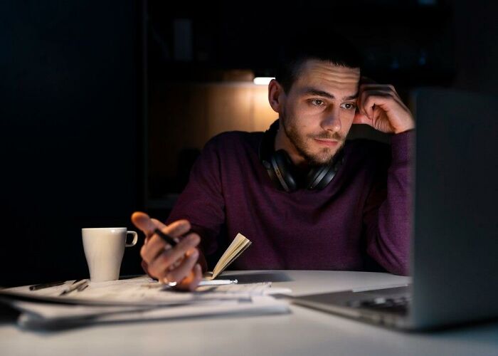 Man working late on a laptop with headphones, papers, and coffee, reflecting on romanticized but awful realities.