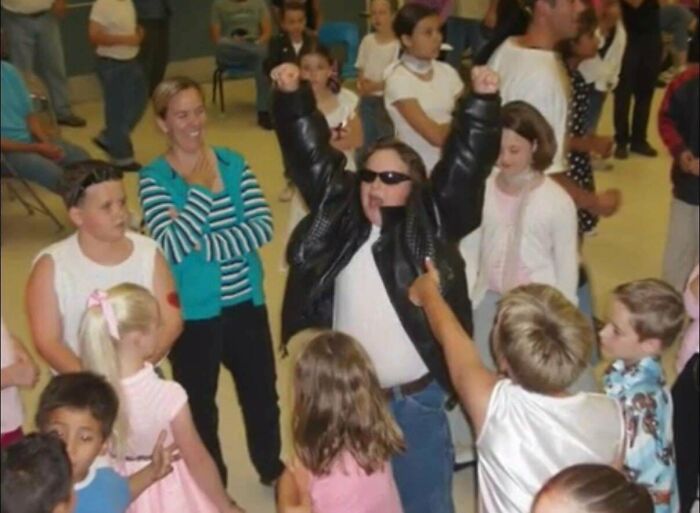 Child in sunglasses and leather jacket raises arms triumphantly, surrounded by kids, during a lively gathering, embodying blunder years.