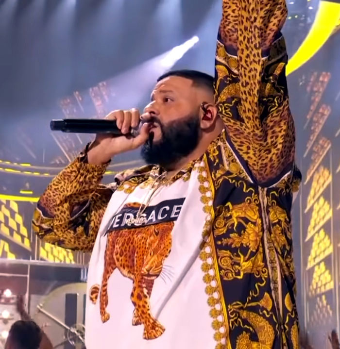 Performer on stage, holding microphone, in vibrant leopard-print outfit, with spotlight in background.