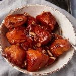 A rustic ceramic bowl filled with glazed sweet potato chunks, garnished with chopped pecans. A spoon rests inside the bowl, and the dish is set against a light-colored fabric backdrop.