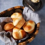 A basket filled with freshly baked golden brown rolls, lined with a light gray cloth. In the background, there is a small dish of butter with a knife on a dark plate. The setting is on a textured blue surface.