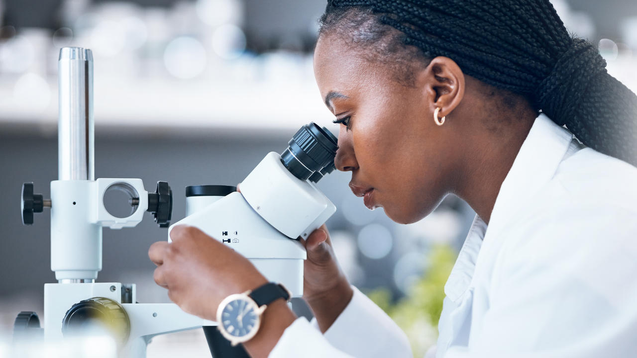 A dark-skinned woman researcher with long, dark, braided hair wearing a white lab coat looks into a microscope while working in a laboratory.