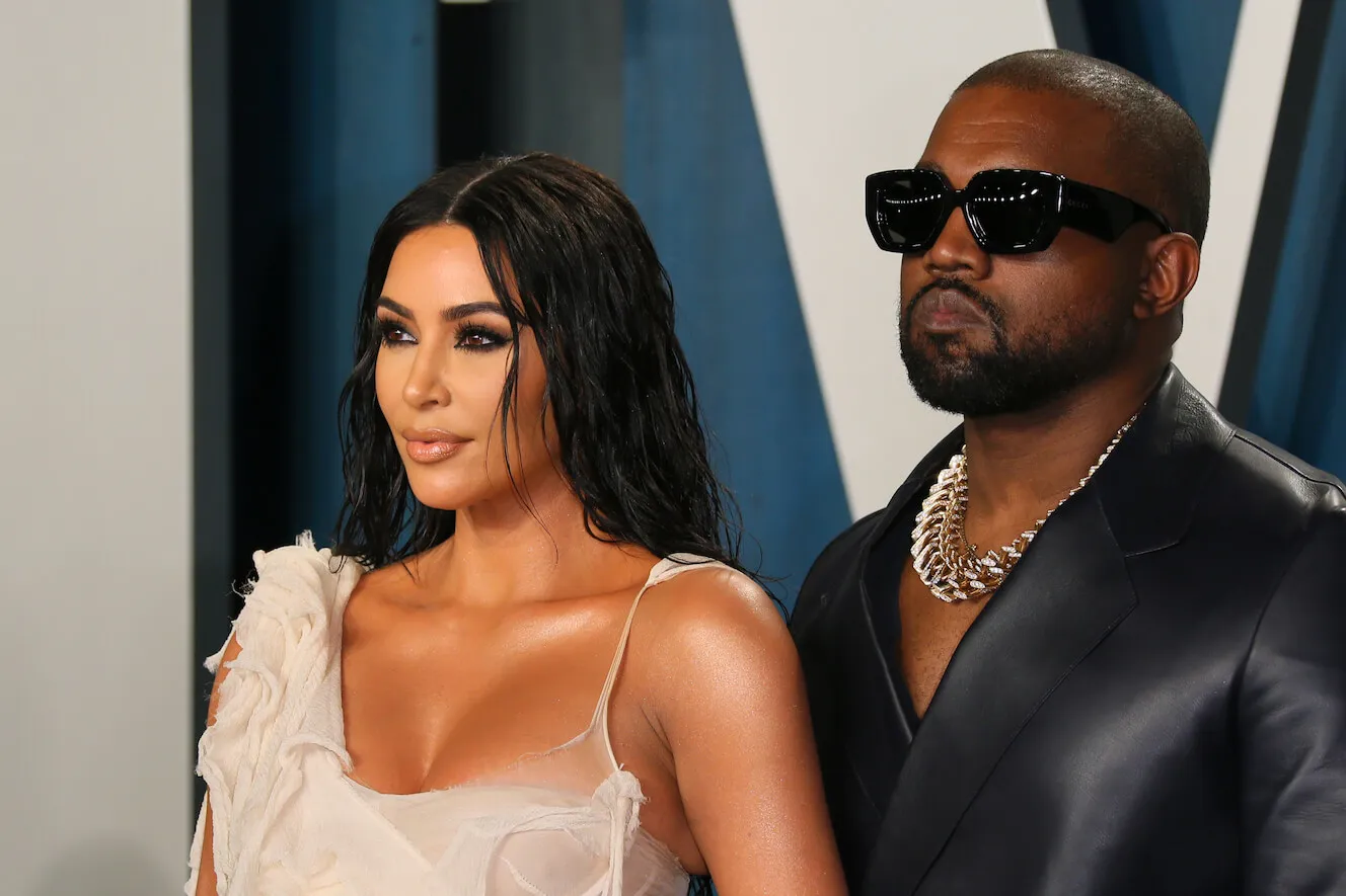 Kim Kardashian and Kanye West posing at the 2020 Vanity Fair Oscar party. Kardashian is wearing a one-shoulder nude dress and West is wearing a black jacket with matching sunglasses.