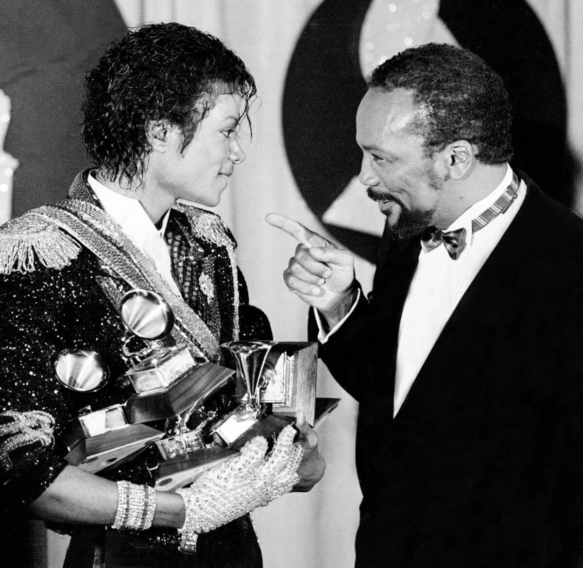 A black and white picture of Michael Jackson holding Grammys and standing with Quincy Jones. Jones points at Jackson.