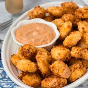 A plate of golden-brown, crispy Air Fryer Popcorn Shrimp is served alongside a bowl of creamy dipping sauce. The shrimp are arranged in an oval dish with a fork visible in the background, and part of a blue and white patterned cloth napkin showing to the side.