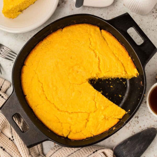 A round pan of golden-brown cornbread with one slice missing sits invitingly in a black cast-iron skillet on a light-colored surface. Nearby are utensils, a beige towel, and a white plate ready to accompany your morning coffee delight.