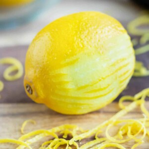 A lemon that has been partially zested on a cutting board with lemon zest around it.