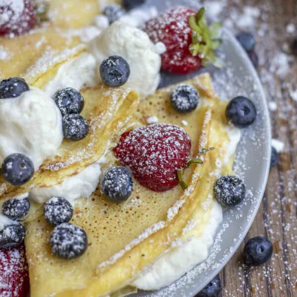 side view of three sourdough crepes with whipped cream filling, blueberries and strawberries on a gray plate on a wood table.