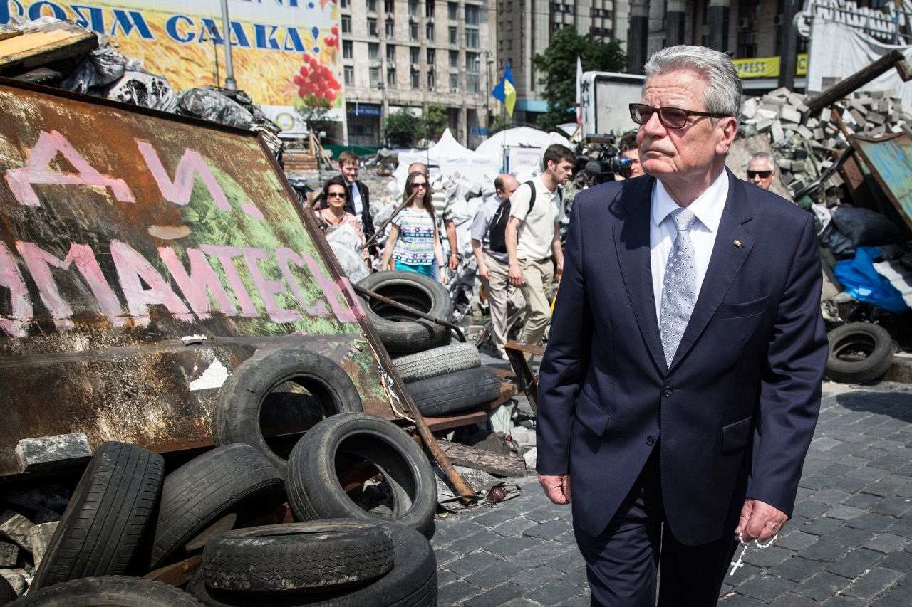 Farbiges Foto: Bundespräsident Joachim Gauck mit Sonnenbrille und Rosenkranz in der linken Hand. Er geht vor Straßenbarrikaden, die unter anderem aus Reifen, Schildern und aufgeschütteten Ziegeln bestehen und teils mit Graffitis in kyrillischen Schriftzeichen besprüht sind. Im Hintergrund mehrere Personen.