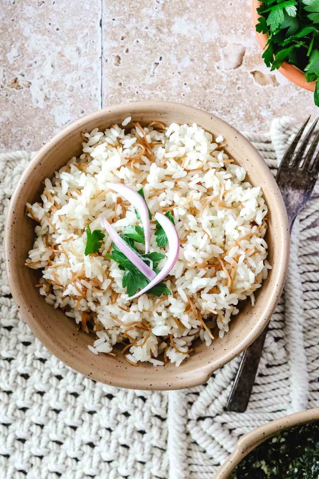 Egyptian rice with vermicelli with sliced red onions and cilantro on top with a fork beside it on a woven background and a bowl of spinach and a bowl of cilantro on a pink tiled background