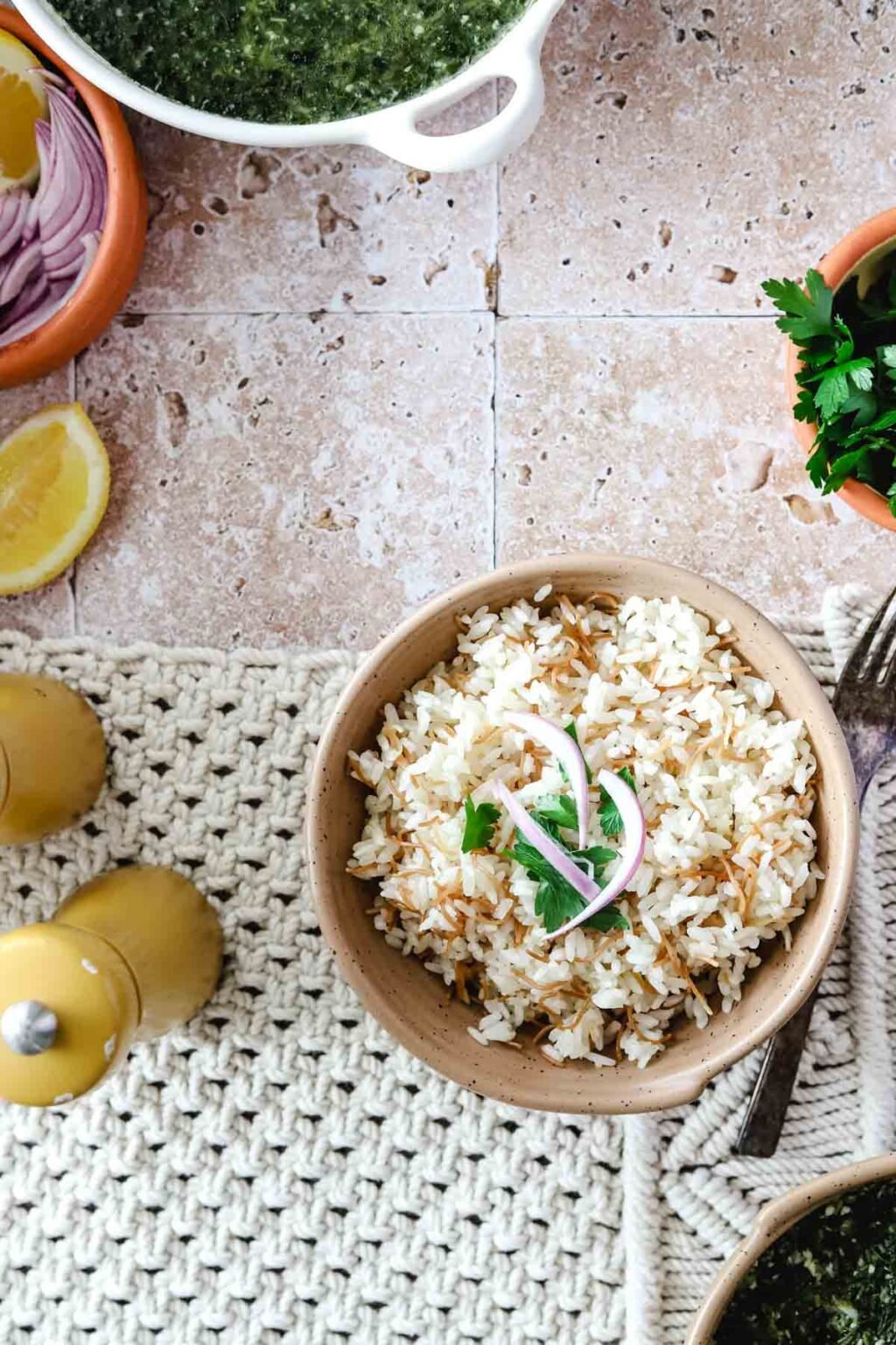 Cooked Egyptian rice and vermicelli in a beige bowl with slices of red onion and cilantro on top on a woven background and a fork beside the bowl of rice, with salt and pepper shakers, wedge of lemon, bowl of onions and bowl of spinach and a bowl of cilantro on a pink tiled background