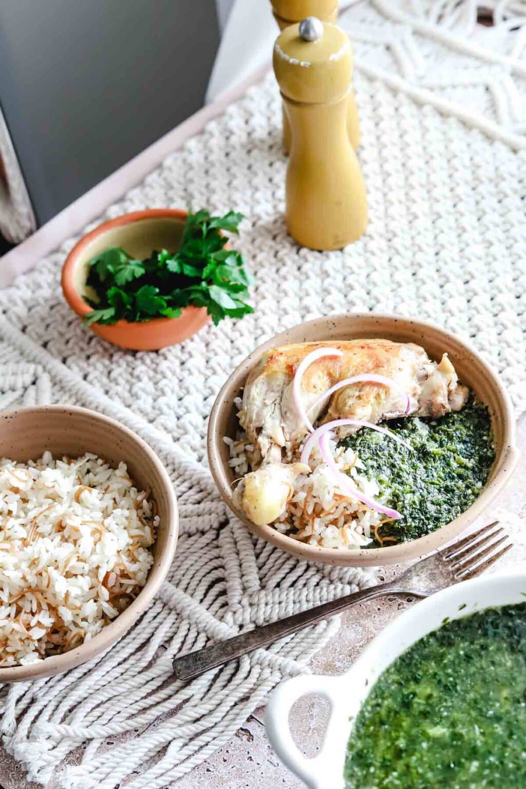 a beige bowl with rice, molokhia, cooked chicken and sliced red onions with a fork beside it on a woven background with molokhia in a white bowl in the corner, rice in a beige bowl in the bottom corner and cilantro in a bowl along with salt and pepper shakers