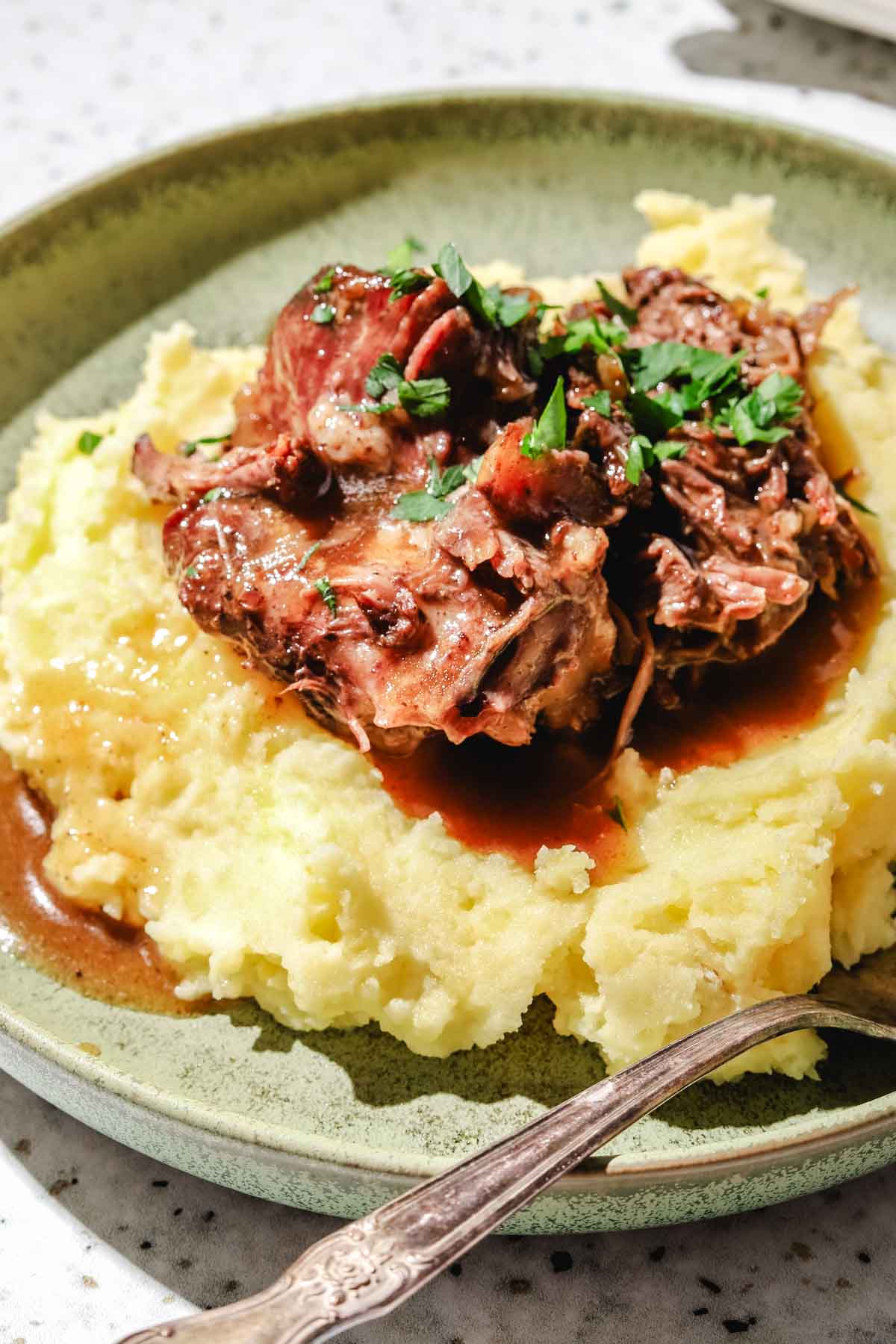 shredded chuck roast served on top of mashed potatoes with the chuck roast juices poured on top with parsley and a fork on the side on a light green plate on a speckled backdrop