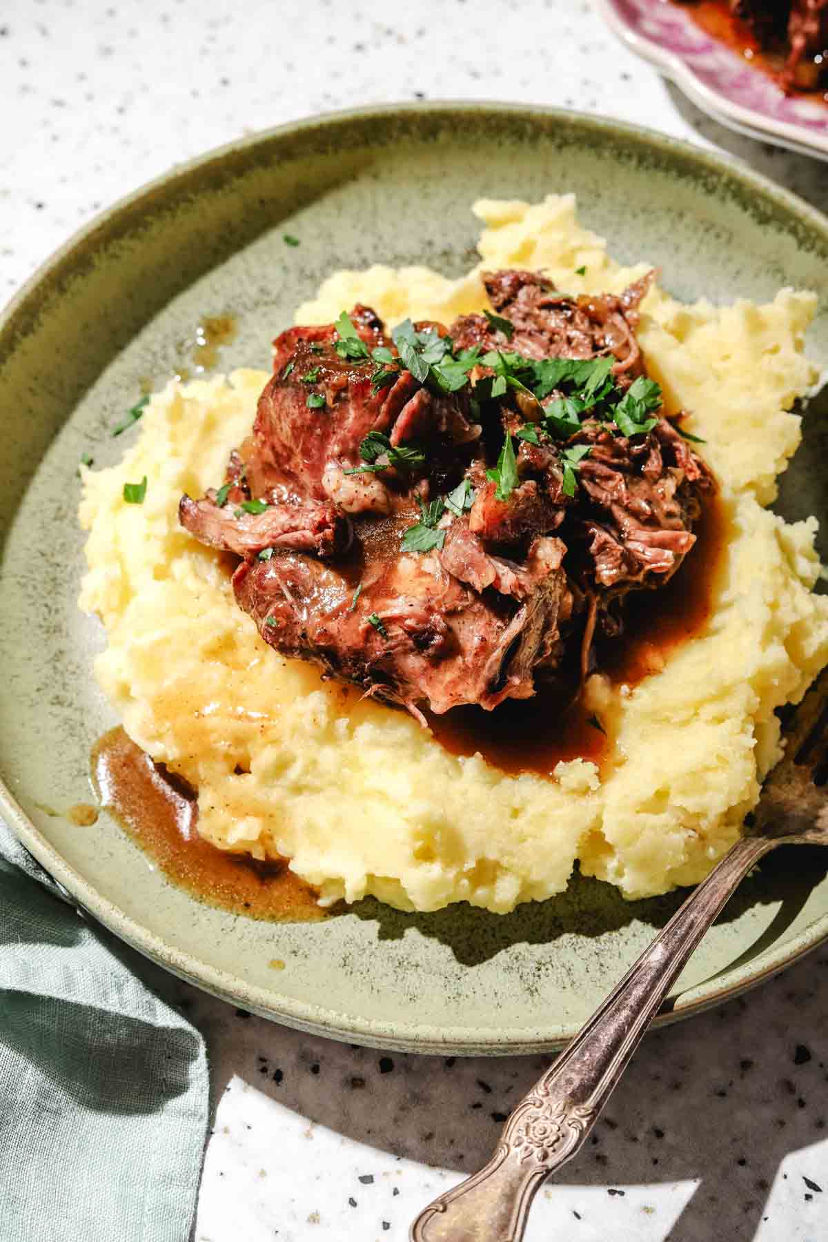 shredded chuck roast served on top of mashed potatoes with the chuck roast juices poured on top with parsley and a fork on the side on a light green plate on a speckled backdrop with a pink edged platter with shredded chuck roast and a green tea towel beside the plate