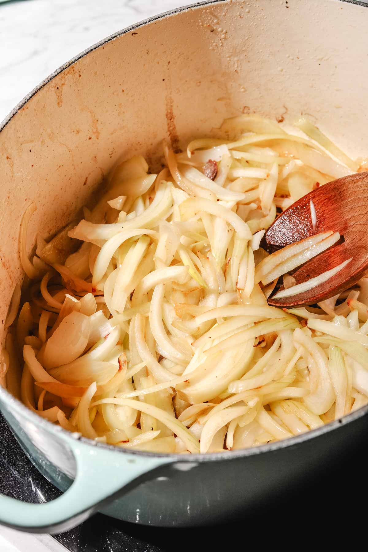 Sliced white onions being sauteed in an oval light mint pot with a wooden spoon on a light marble background