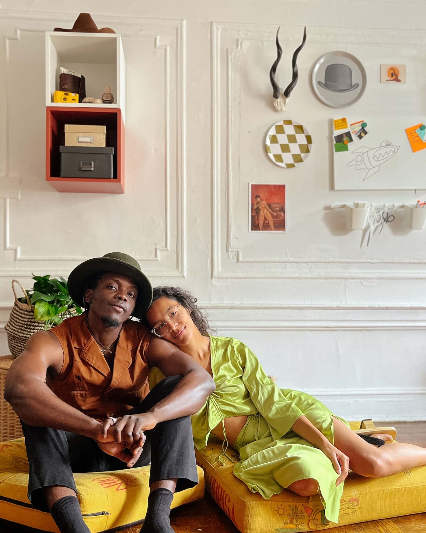 A couple sit together on the floor in front of a wall adorned with artwork and ornaments.