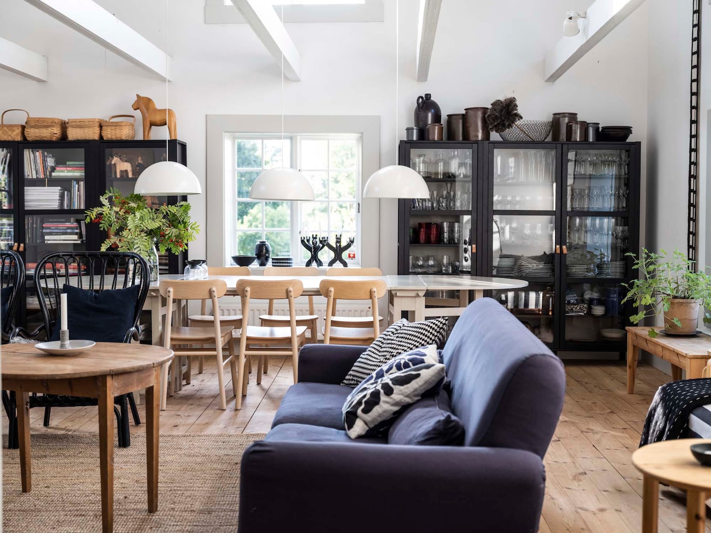 A livingroom with a blue sofa, light wood dining table and chairs and two black wooden display cabinets. Two white pendant lights are hung above the dining table.