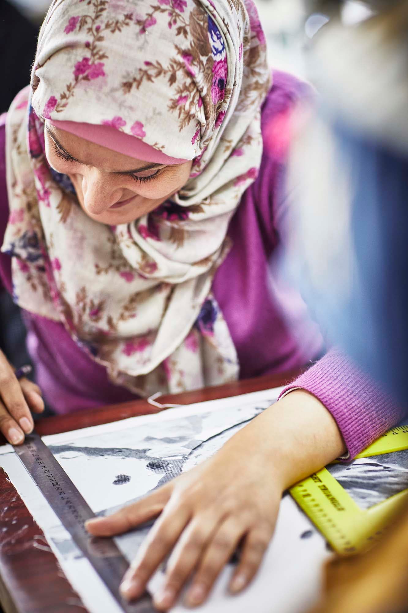 A woman wearing a floral hijab measures a drawing with a ruler. She smiles as she works.