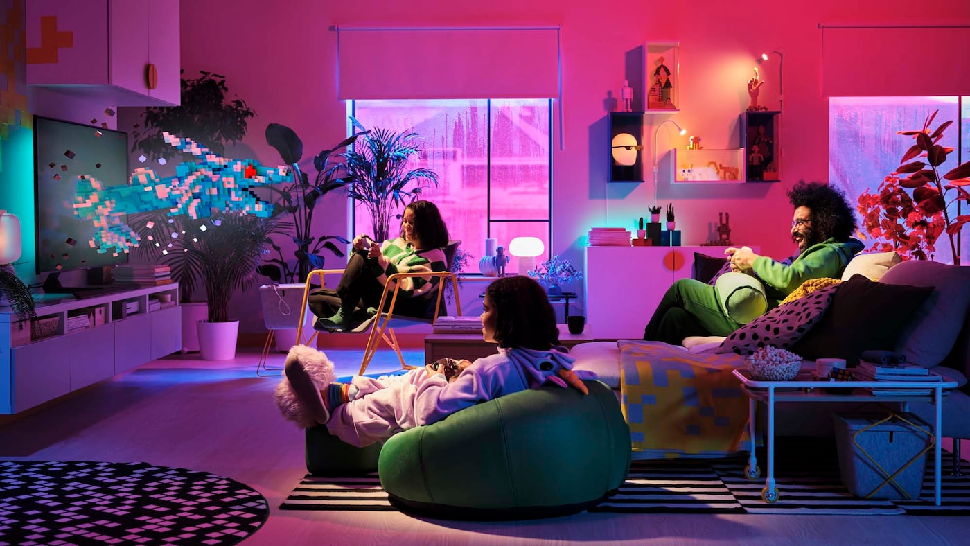 An adult and two teens gaming in a pink fluorescent-lit living room, furnished with products from the BRÄNNBOLL gaming collection.