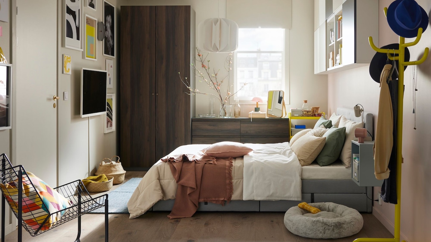 Wide angle view of a bedroom styled in pale neutrals with dark wood cabinets, and accents of bright yellow and soft pinks.