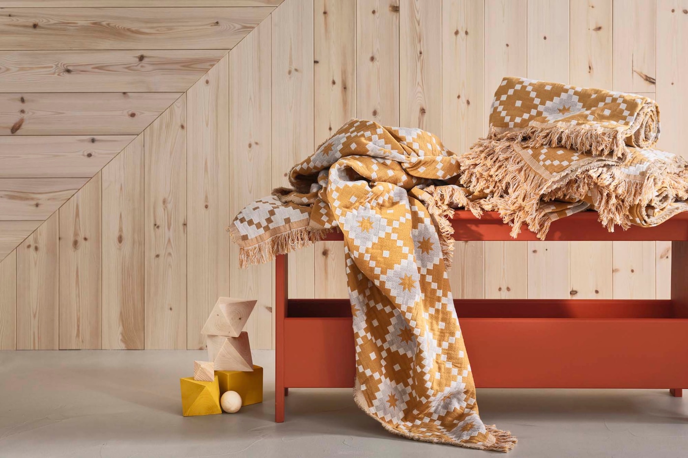 A terracotta coloured PERJOHAN bench stands against a wood wall backdrop. On the bench are throws stacked and falling off. Beside the bench is a yellow lantern.