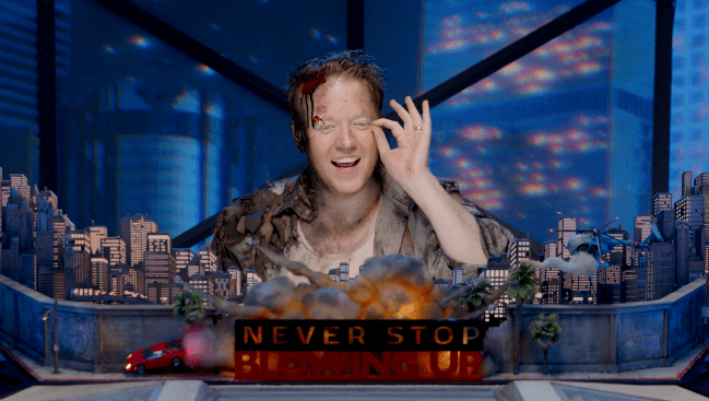 Still of Brennan Lee Mulligan as the game master of 'Never Stop Blowing Up' sitting in front of a DM screen shaped like a city skyline with a car and smoke behind a title card that reads 'Never Stop Blowing Up.' 