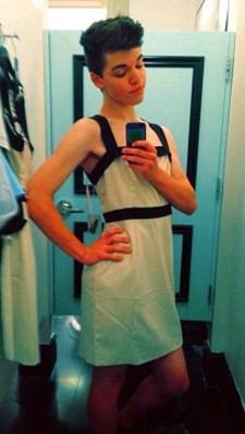 Teenaged, white, transgirl with dark hair wearing a white dress and posing in front of a mirror, taking a photograph of himself using a camera phone.