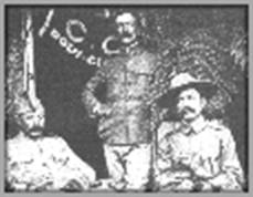 Photograph of three men at a boy scout event circa 1910.  The man seated on the left is unidentified, Burnham is in the middle, standing, and Baden-Powell is on the right, seated.  There is a table in front of the men.  Baden-Powell is wearing his stetson hat, Burnham has no hat, and man on the left has a modest hat.  Behind Burnham and to his right is a flag partially opened.