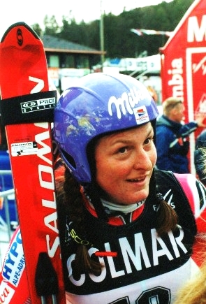 Janica Kostelić standing and holding skis, wearing a helmet and dressed in a ski racing suit.