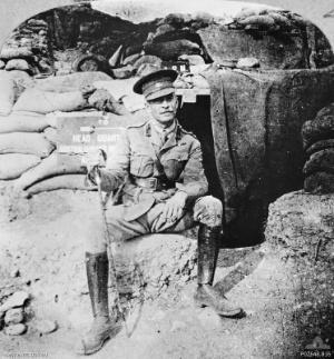 Soldier wearing Sam Browne belt and peaked cap, with a walking stick, sitting in front of a sandbagged doorway half covered by a tarpaulin. A sign says: "headquarters".