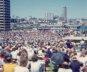 A large crowd. Far away is a platform, and a banner reading SHAME FRASER SHAME.