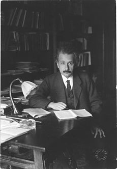 Einstein, sitting at a table, looks up from the papers he is reading and into the camera.