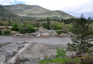 A muddy river flows through a gap between the ruins of a dam on either shore. A wooded hill or mountain rises in the background.