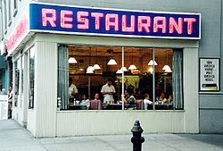 The exterior of a restaurant at the corner of a street. Through the windows a waiter can be seen taking orders. Above the windows is the word "Restaurant" in big pink letters.