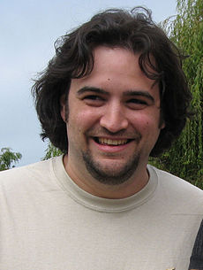 Causcasian man with medium-length dark brown hair and a beard smiles for a camera. The man is wearing a gray T-shirt with an image of evolution from a monkey to a pirate