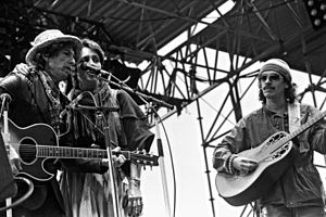 On stage outdoors, Dylan in a leather jacket and iconic porkpie hat & scarf, Baez wears a peasant skirt, sarape, tassled scarf around neck, her arm around Dylan's shoulder. Santana in workman's shirt & knit cap.