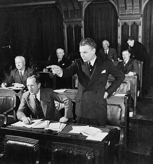 Diefenbaker, standing in a legislative chamber, dramatically points in front of him. His hair is greying, and he appears much the way he will as Prime Minister.