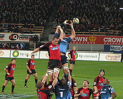 Two teams of men, one dressed in red and the other in blue, standing on a grass field in what appears to be a stadium. The teams are competing to reach a ball several feet above head-height. To achieve this, one man from each team is raised into the air by two of his team-mates; these men's knees are above their team-mates heads.