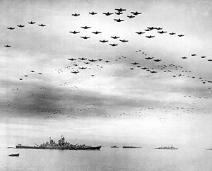 Black and white photo showing a large number of aircraft flying in formation over several World War II-era warships