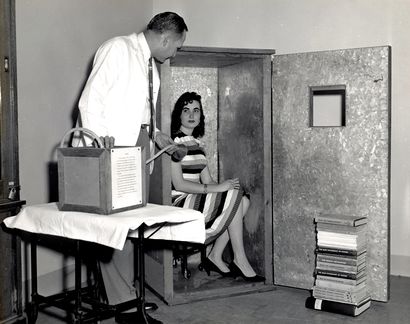 photograph of a woman, sitting on a chair which is in a large, wardrobe-sized  box, the open door of which has a small window. A man stands nearby, holding a breathing apparatus.