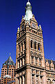 Ground-level view of a large, brick tower with several architectural niches and columns and a tapering, copper roof topped with a spire; a clock are visible on the tower near its roofline.